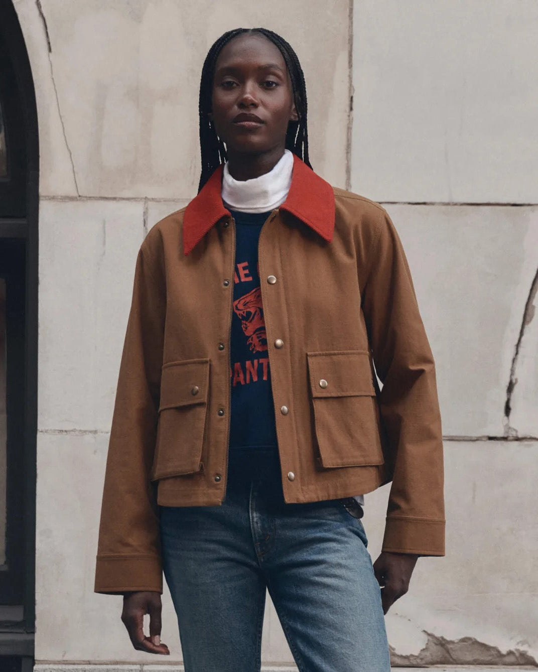 A model wearing a brown cropped canvas jacket with a contrasting red collar, layered over a white turtleneck, a navy graphic tee, and blue jeans, standing against a light-colored stone wall.
