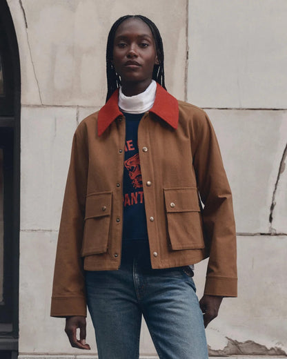 A model wearing a brown cropped canvas jacket with a contrasting red collar, layered over a white turtleneck, a navy graphic tee, and blue jeans, standing against a light-colored stone wall.